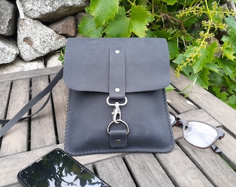May include: A black leather crossbody bag with a silver clasp and a stitched edge. The bag is sitting on a wooden surface with a cell phone and glasses.