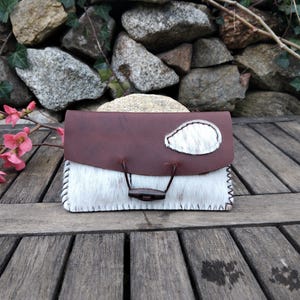 Può includere: Un piccolo portafoglio in pelle marrone con una patta in pelliccia bianca e una chiusura a cinturino in pelle marrone. Il portafoglio è appoggiato su una superficie in legno.