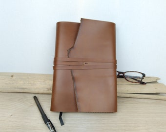 May include: A brown leather journal with a strap closure. The journal is closed and lying on a wooden surface with a pen and glasses nearby.