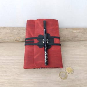 Pode incluir: Diário de couro vermelho com fecho elástico preto e design de flor preta. Uma caneta preta está presa na banda elástica.