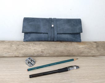 May include: A blue leather pencil case with a flap closure. The case is sitting on a piece of driftwood. There is a pencil sharpener, a pencil, and a pen on a wooden surface.