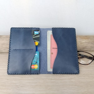 Puede incluir: Cartera de cuero azul marino abierta con ranuras para tarjetas y un bolsillo para documentos. La cartera está abierta, revelando tarjetas de crédito y un documento rosa. Los bordes tienen un patrón de costura decorativo. Gafas de carey descansan sobre la mesa.