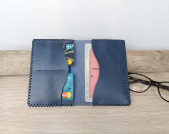 May include: Open navy blue leather wallet with card slots and a document pocket. The wallet is open, revealing credit cards and a pink document. The edges have a decorative stitched pattern. Tortoiseshell eyeglasses rest on the table.