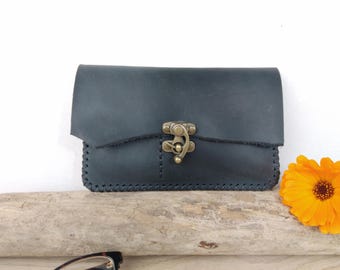 May include: A dark blue leather clutch with a brass clasp. The edges are hand-stitched. A bright orange flower is in the background. The clutch is approximately 7 inches wide and 4 inches tall.