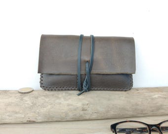 May include: A handcrafted, brown leather glasses case with a flap closure and a dark blue leather tie. The case is stitched along the edges and rests on a piece of driftwood. A pair of eyeglasses is visible in the foreground.