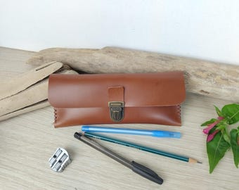 May include: A rectangular brown leather pencil case featuring a brass-colored metal push-lock closure and visible stitching along the sides. The case is accompanied by a silver metal pencil sharpener, a black pen, and two pencils.