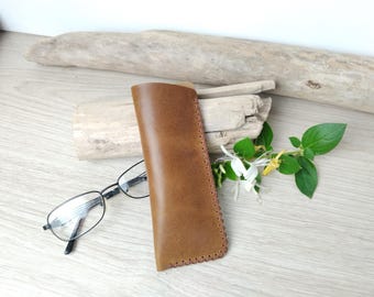 May include: A brown leather glasses case with a stitched edge, next to a pair of black-framed eyeglasses. The case is resting on a light-colored wooden surface, with a piece of driftwood and greenery in the background.