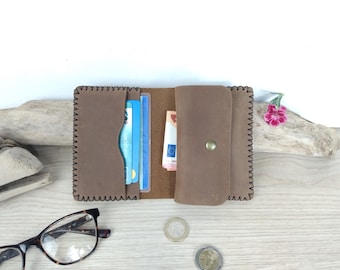 May include: A brown leather wallet featuring visible whipstitch detailing along the edges. The wallet is open, revealing interior card slots containing blue and white cards, a central compartment holding paper currency, and a flap with a metal snap.
