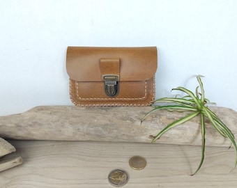 May include: A tan leather pouch featuring a rectangular flap, a bronze-toned metal push-lock closure, and visible whip-stitching along the bottom edges. The pouch has a smooth finish and a structured design with a centered front clasp mechanism.