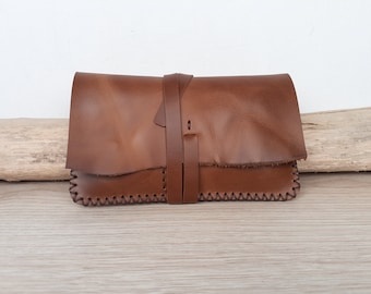 May include: A brown leather clutch with a strap closure. The clutch has a stitched edge and a flap closure.