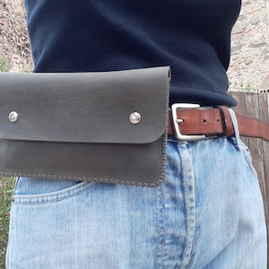 May include: A dark gray leather belt bag with a stitched edge and silver metal snaps. The bag is worn around the waist of a person wearing blue jeans and a brown leather belt.