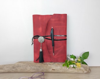 May include: A red leather journal with a black pen, secured by a black leather strap and a decorative silver button. A black tassel hangs from the button. The journal is on a piece of driftwood.