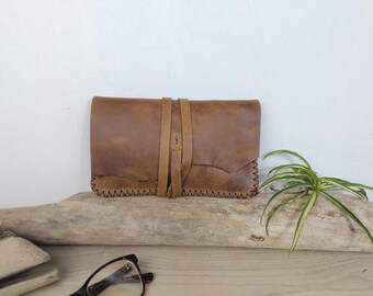 May include: A rectangular brown leather pouch featuring a wrap-around tie closure and decorative whipstitch detailing along the bottom edge. The leather has a smooth, matte finish with natural variations in color and texture across the surface.