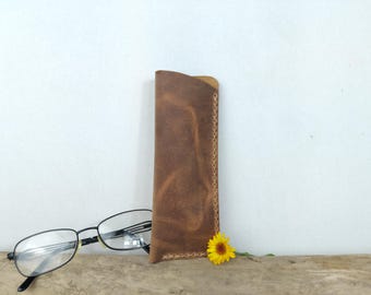 May include: A brown leather glasses case with a stitched edge. The case is standing upright next to a pair of black-framed glasses and a yellow flower. The case is made of a textured leather material.