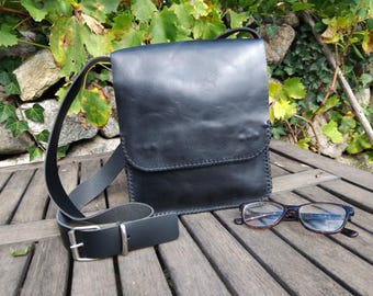 May include: Black leather crossbody bag with a flap closure and stitched edges. A matching black belt with a silver buckle and a pair of eyeglasses rest on a wooden surface. The background features foliage and a stone wall.