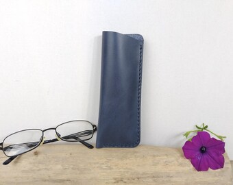 May include: A navy blue leather glasses case with hand-stitched edges stands upright next to a pair of black-framed eyeglasses. A vibrant purple flower with a dark center rests on a wooden surface.