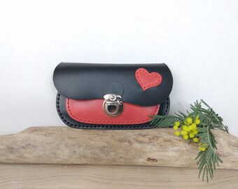 May include: A small, black leather purse with a red heart applique and a red bottom. The purse has a silver clasp and is displayed on a piece of driftwood. A sprig of yellow flowers and green leaves is in the background.