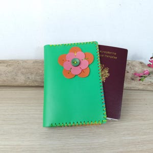 Op de afbeelding: Een groene paspoorthouder met een bloemdesign van oranje en roze cirkels en een groene knoop. Er ligt een bordeauxrode paspoort naast. De houder is met gele draad met de hand gestikt.