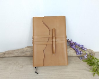 May include: A tan leather journal with a wrap-around strap closure and a matching tan pen secured in a leather loop on the front cover. The journal features a vertical, uneven, torn-style edge detail on the left side of the front cover.