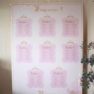 May include: A large, pink and gold wedding seating chart with eight tables, each with a crown design and guest names. The text "&eacute;tait une fois..." is at the top, with "Be our guest..." at the bottom. The words "LES MOTS inspirants" are at the bottom left.