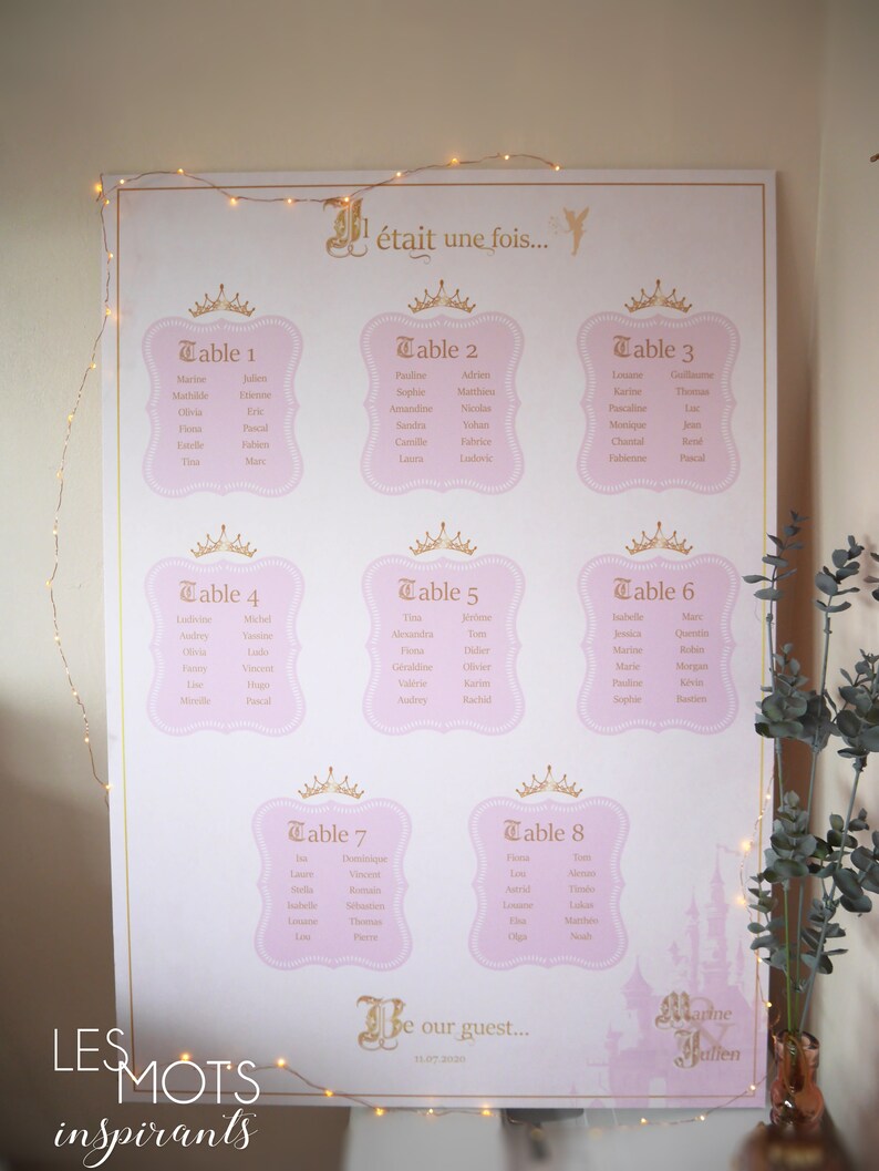 May include: A large, pink and gold wedding seating chart with eight tables, each with a crown design and guest names. The text "&eacute;tait une fois..." is at the top, with "Be our guest..." at the bottom. The words "LES MOTS inspirants" are at the bottom left.
