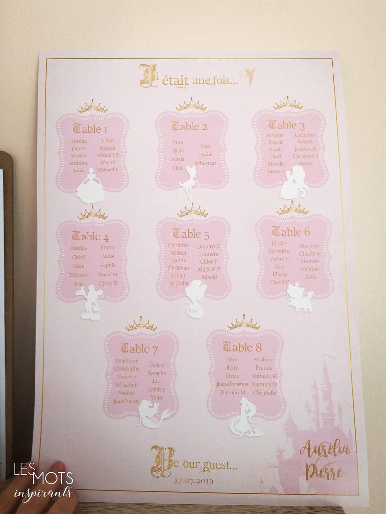 May include: A pink wedding seating chart with gold accents and a fairytale theme. The chart features eight tables with guest names, decorative crowns, and Disney character cutouts. The text "Be our guest..." is also visible.