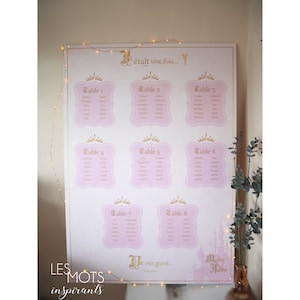 May include: A large, pink and gold wedding seating chart with eight tables listed. Each table is in a pink frame with a gold crown. The text "&Eacute;tait une fois..." is at the top, and "Be our guest..." is at the bottom.