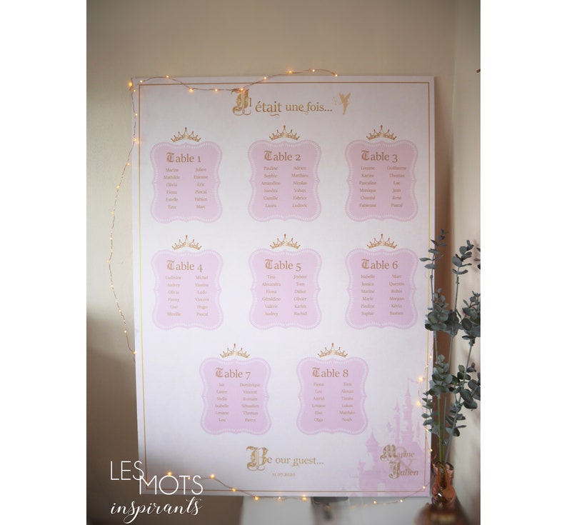 May include: A large, pink and gold wedding seating chart with eight tables listed. Each table is in a pink frame with a gold crown. The text "&Eacute;tait une fois..." is at the top, and "Be our guest..." is at the bottom.