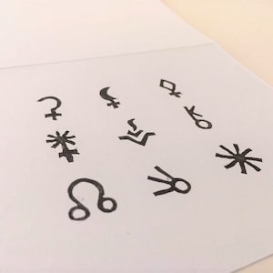 以下が含まれることがあります： 白いカードに9つの黒い占星術のシンボルが描かれています。シンボルには、太陽、月、水星、金星、火星、木星、土星、天王星、海王星が含まれています。