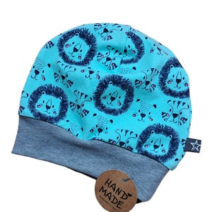Könnte beinhalten: Eine türkisfarbene Baby-Beanie-Mütze mit einem sich wiederholenden Muster aus Löwengesichtern und anderen Tierillustrationen. Der Rand ist grau, und ein kleines Etikett mit einem Stern ist angebracht. Ein braunes Etikett mit "HAND MADE" ist ebenfalls angebracht.