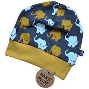 Könnte beinhalten: Eine marineblaue Beanie-Mütze mit einem senfgelben Rand. Die Mütze ist mit hellblauen und senfgelben Elefanten gemustert. Ein kleines Etikett mit der Aufschrift "HAND MADE" ist angebracht.