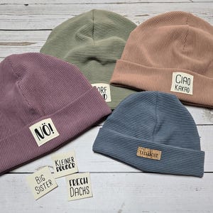 Könnte beinhalten: Eine Sammlung von vier gerippten Strickmützen in Lila, Grün, Rosa und Blau. Jede Mütze hat ein rechteckiges Etikett mit Text. Zusätzliche Etiketten mit Text sind darunter verstreut.