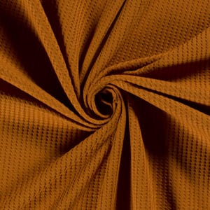 Könnte beinhalten: Nahaufnahme einer Wirbel aus strukturiertem, goldbraunem Stoff. Das Material hat ein waffelartiges Muster mit vertikalen Linien und kleinen Quadraten. Der Stoff ist dicht gewebt und wirkt weich. Die Farbe ist ein warmer, erdiger Ton.