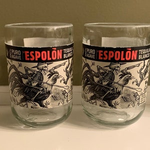 May include: Two clear glass tumblers with a decorative label featuring a skeleton riding a rooster. The label has the text "ESPOLON TEQUILA BLANCO" in red and white. The glasses are likely for serving tequila.