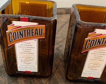 May include: Two square-shaped, amber-colored glasses with the "COINTREAU" logo in orange and white. The glasses are made from recycled liquor bottles, with the original labels still attached. The glasses are sitting on a wooden surface.