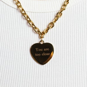 Op de afbeelding: Goudkleurige ketting met een hartvormige hanger. De hanger heeft de gegraveerde tekst "You are too close". Het woord "hope" is op de nek getatoe&euml;erd.