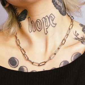 Puede incluir: Una persona con el pelo corto rubio y varios tatuajes, incluyendo una mariposa y la palabra "esperanza" en el cuello. Lleva un top negro de tirantes y un collar de cadena plateada. La persona tiene varios piercings en la cara y las orejas.