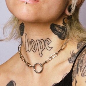 Może przedstawiać: Osoba z krótkimi blond włosami i wieloma kolczykami w twarzy. Ma tatuaż z napisem "nadzieja" na szyi i kilka innych tatuaży na ramionach i barkach.