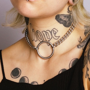 Puede incluir: Un collar de cadena plateada con un gran colgante circular. La palabra "hope" está tatuada en el cuello de la persona. La persona tiene múltiples piercings y tatuajes faciales, incluyendo una mariposa y fases lunares.