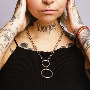 May include: A person with short blonde hair and multiple tattoos, including the word "hope" on their neck, is wearing a black tank top and a silver chain necklace with two large rings. They have several piercings on their face and ears.