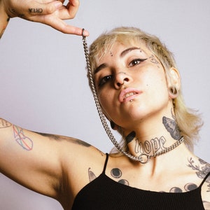 Puede incluir: Un collar gargantilla de cadena plateada con una cadena a juego que se extiende hacia arriba. La modelo lleva una camiseta sin mangas negra de canalé. Tatuajes adornan los brazos y el cuello. La palabra "hope" está tatuada en el cuello.