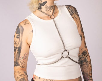 May include: A person with short blonde hair and numerous tattoos is wearing a white vest top with a silver chain body harness. The word "hope" is tattooed on their neck.