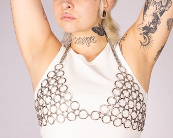 May include: A woman with short blonde hair and tattoos is wearing a white tank top with a silver chainmail top. The chainmail top is made of interlocking metal rings. The woman has the word "hope" tattooed on her neck.