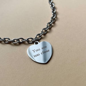 Op de afbeelding: Zilverkleurige ketting met een hartvormige hanger met de gegraveerde tekst "You are too close". De ketting bestaat uit ovale schakels. Een sieraad.