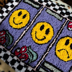 Puede incluir: Una alfombra morada con un borde a cuadros presenta tres caras sonrientes amarillas, dos con expresiones felices y una con una expresi&oacute;n triste. La alfombra tambi&eacute;n tiene dise&ntilde;os florales coloridos.