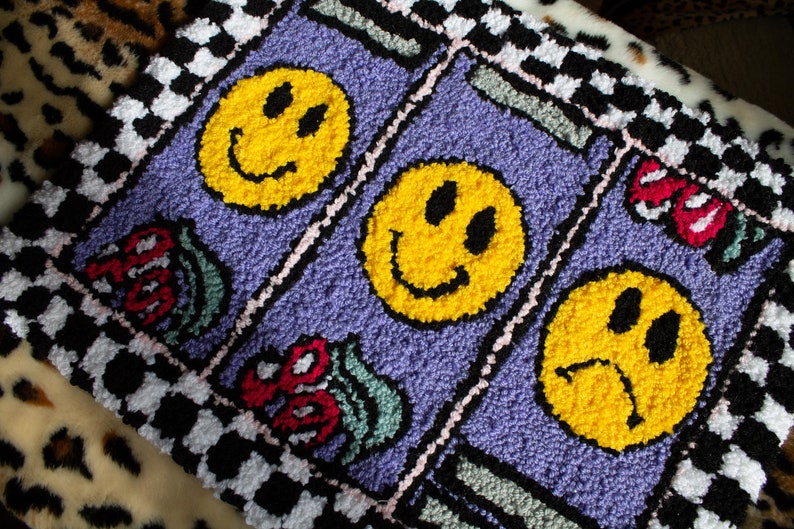 Puede incluir: Una alfombra morada con un borde a cuadros presenta tres caras sonrientes amarillas, dos con expresiones felices y una con una expresi&oacute;n triste. La alfombra tambi&eacute;n tiene dise&ntilde;os florales coloridos.