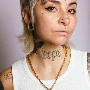 Op de afbeelding: Een gouden ketting met een hartvormige hanger met de tekst "You are too close." De ketting wordt gedragen over een witte tanktop. Het woord "hope" is op de nek getatoe&euml;erd.