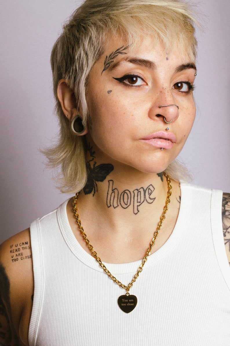 Op de afbeelding: Een gouden ketting met een hartvormige hanger met de tekst "You are too close." De ketting wordt gedragen over een witte tanktop. Het woord "hope" is op de nek getatoe&euml;erd.