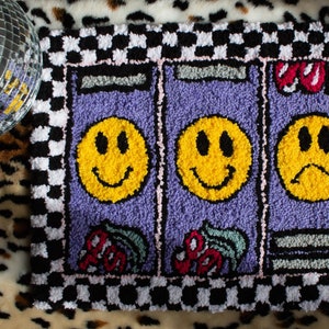 Puede incluir: Una alfombra a cuadros de color morado y negro con tres caras sonrientes amarillas, dos con expresiones felices y una con una expresi&oacute;n triste. La alfombra tiene un borde a cuadros y un borde rosa y blanco.