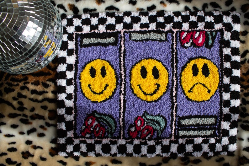 Puede incluir: Una alfombra a cuadros de color morado y negro con tres caras sonrientes amarillas, dos con expresiones felices y una con una expresi&oacute;n triste. La alfombra tiene un borde a cuadros y un borde rosa y blanco.